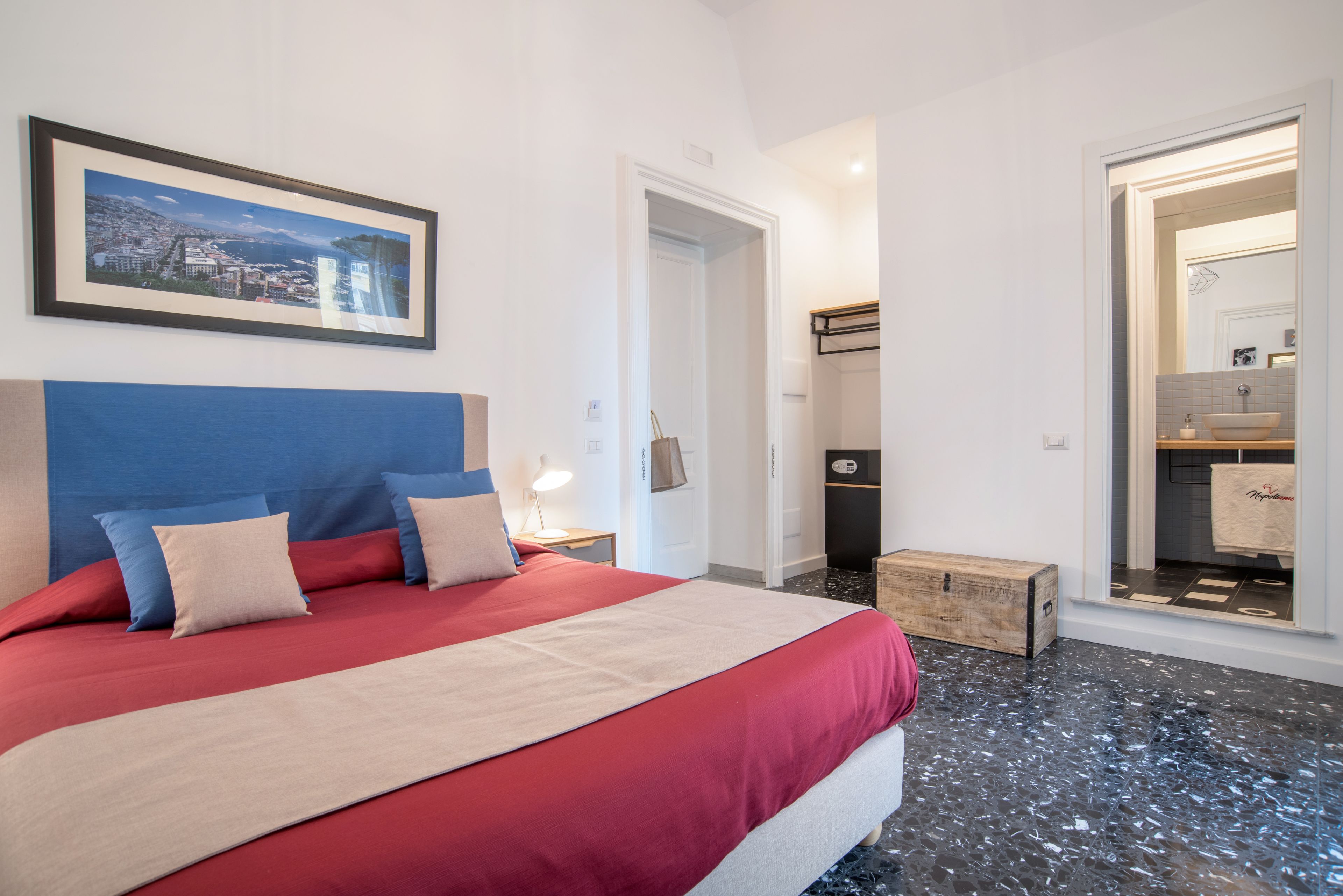A hotel room with a large bed, a framed cityscape picture, a wooden bench, and a bathroom visible through an open door.