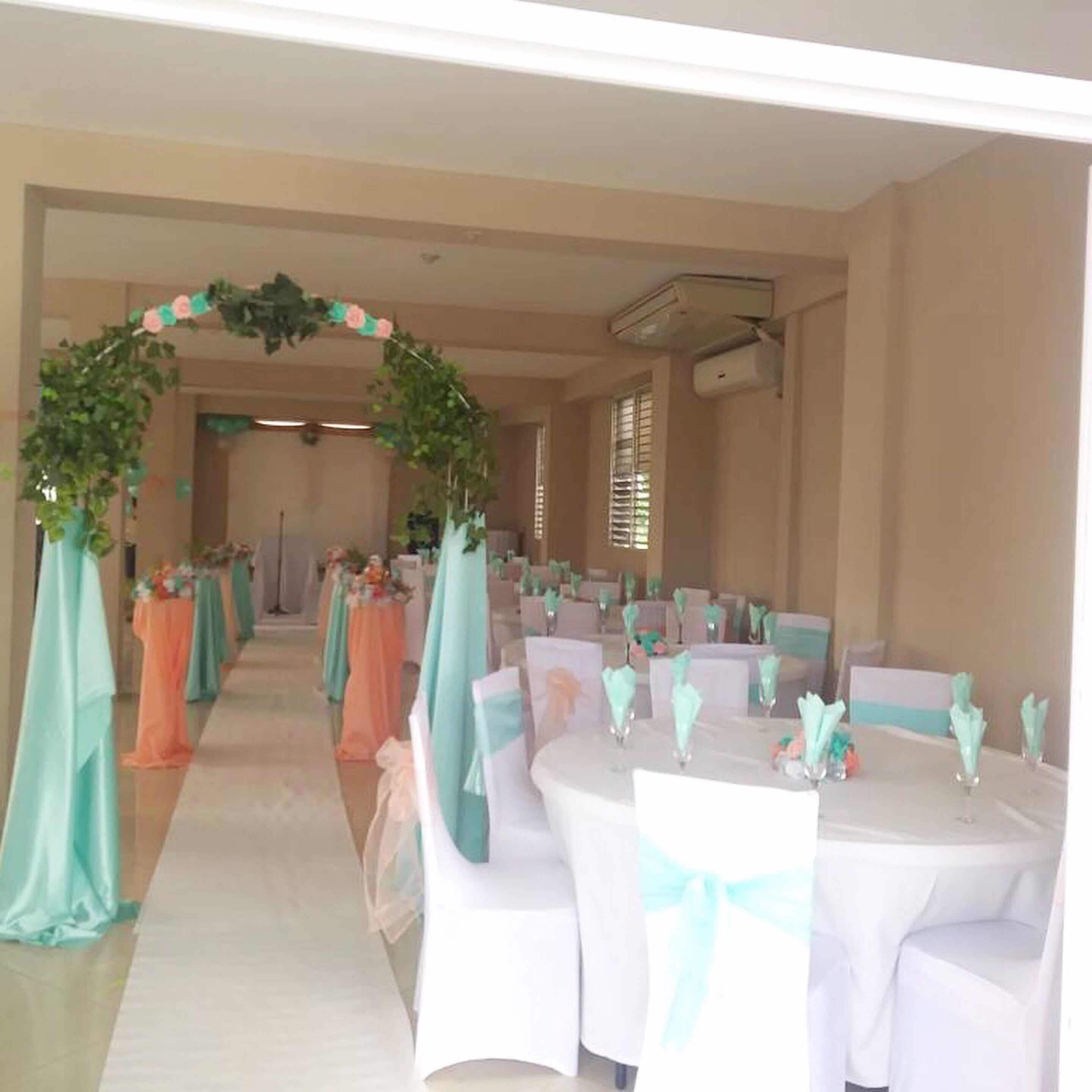 Une salle de banquet avec des tables dressées pour un événement, une arche décorée de fleurs à l’entrée et un climatiseur fixé au mur.