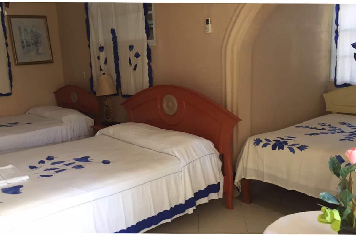 Una habitación con dos camas, cada una con una colcha blanca con motivos florales azules. Una cama tiene un cabecero rojo y la otra, uno amarillo. En la pared hay un cuadro enmarcado, y una lámpara está sobre una mesita de noche.