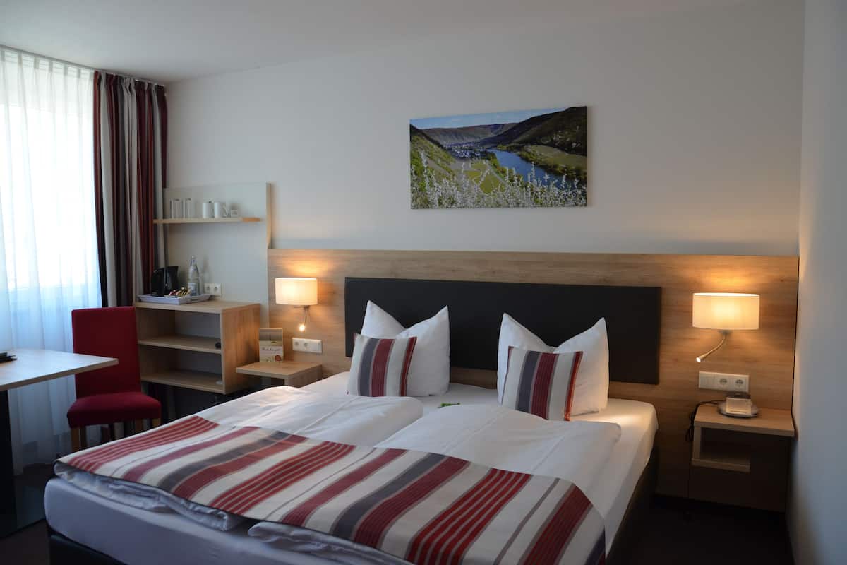 Quarto de hotel com cama, mesas de cabeceira, escrivaninha, cadeira e um quadro na parede.