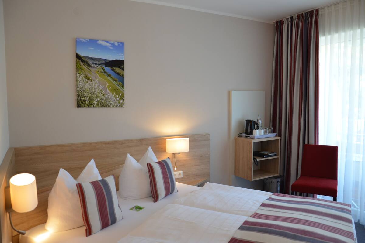 Quarto de hotel com cama, abajures, escrivaninha com cadeira, estante de livros e um quadro na parede.
