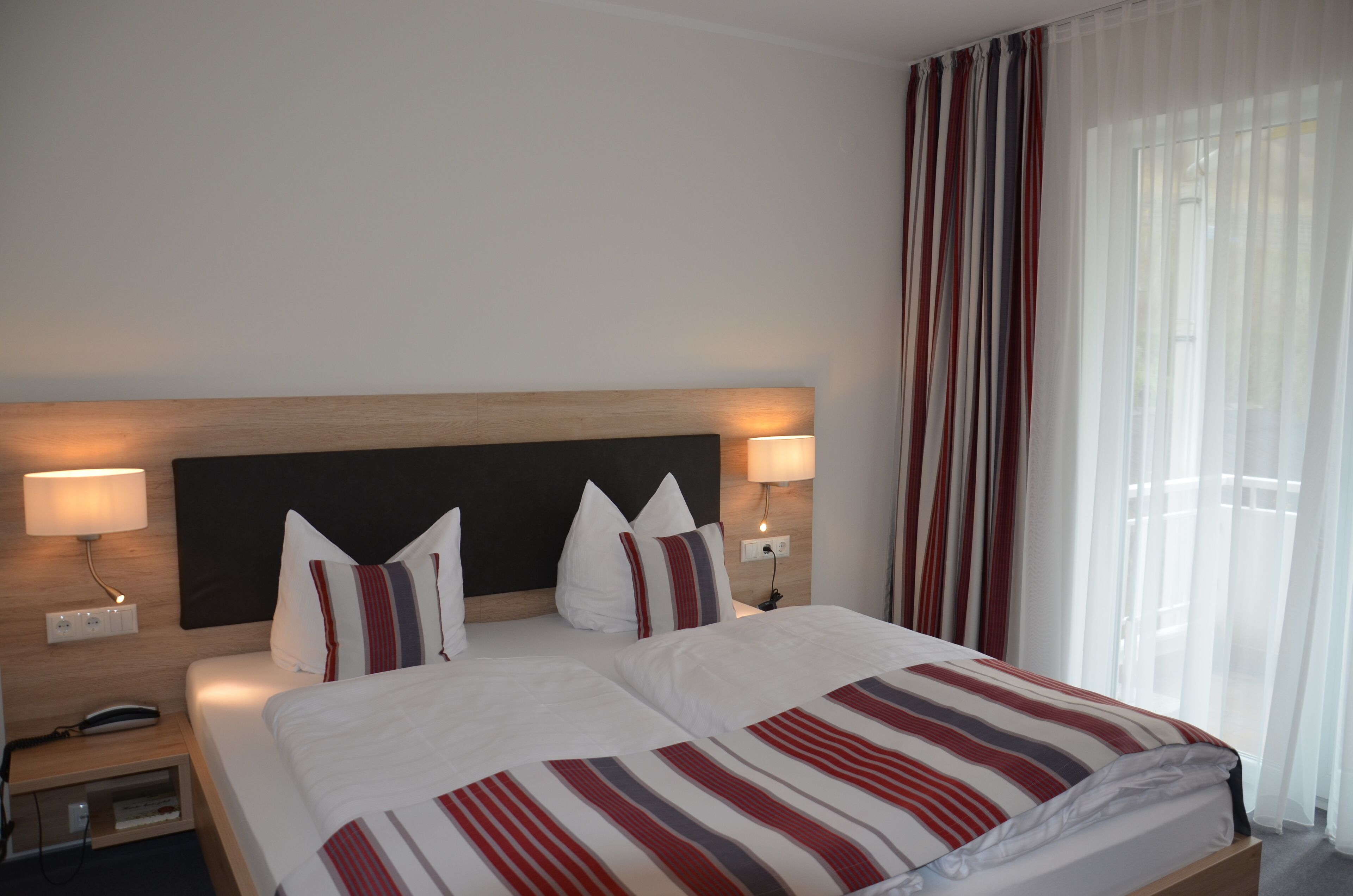 Habitación de hotel con cama doble, dos lámparas de noche, cabecera de madera y ventana con cortinas traslúcidas.