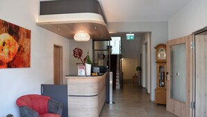 A reception area with a curved desk, a red chair, a large abstract painting, and a wooden cabinet with a clock. - Flair Hotel Am Rosenhügel (Cochem)