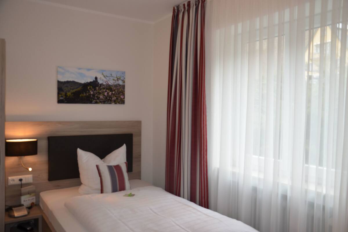 Quarto de hotel com cama, mesa de cabeceira, luminária, quadro na parede e janela com cortinas.