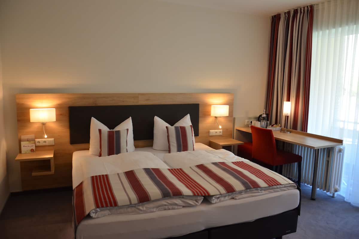 Quarto de hotel com uma cama grande, duas mesas de cabeceira com luminárias, uma escrivaninha com cadeira e vista para o exterior através de cortinas leves.