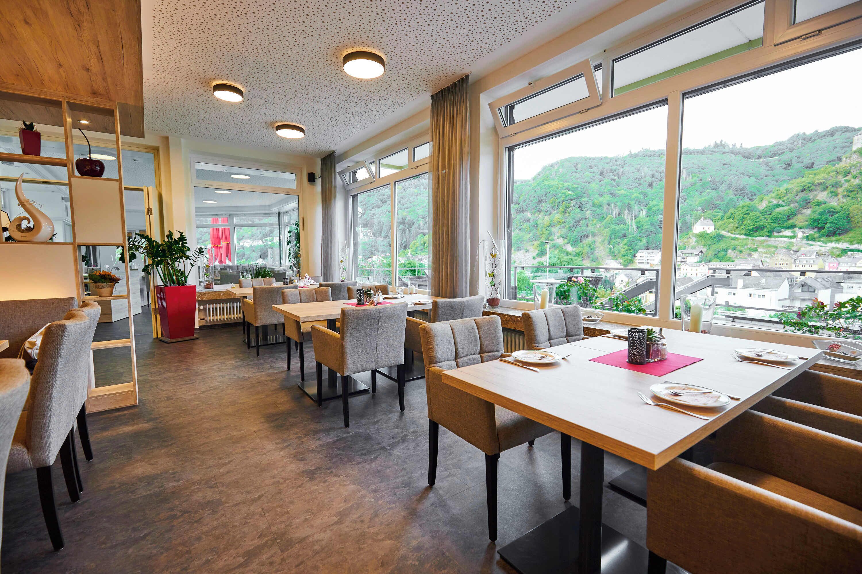 A modern dining area with tables and chairs, large windows with a view of a hilly landscape, and a wooden ceiling.