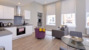 A modern kitchen with white cabinets, a stainless steel range hood, and a built-in oven. There is a purple armchair and a grey sofa in the living area, with a glass coffee table.