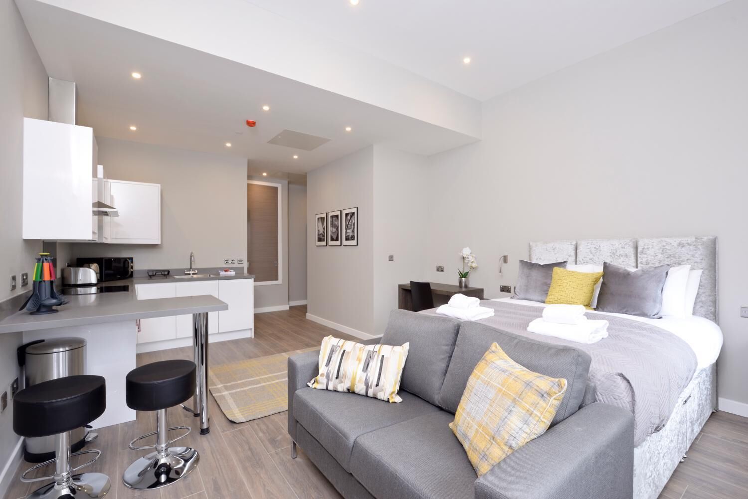 Una habitación de hotel moderna con un sofá gris, una cama con cabecera gris, una pequeña cocina y dos taburetes de bar.