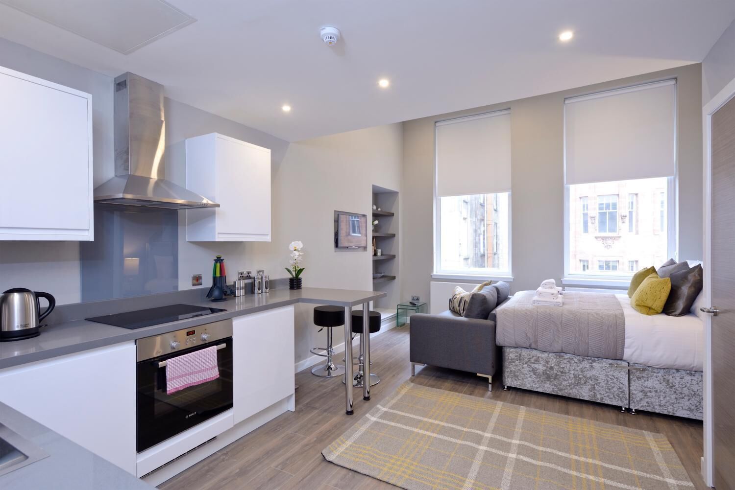 A compact apartment with a kitchenette, a bed with pillows, a small seating area, and a view of the city through large windows.