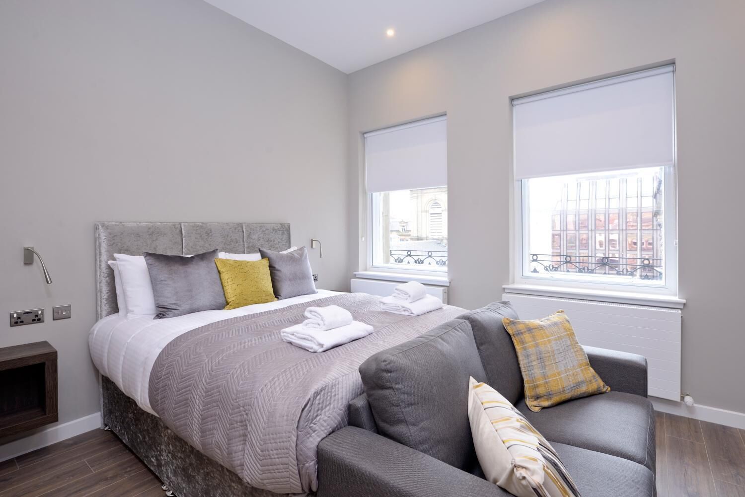 A modern bedroom with a large bed, grey and yellow pillows, a grey sofa, and a view of the city through the windows.