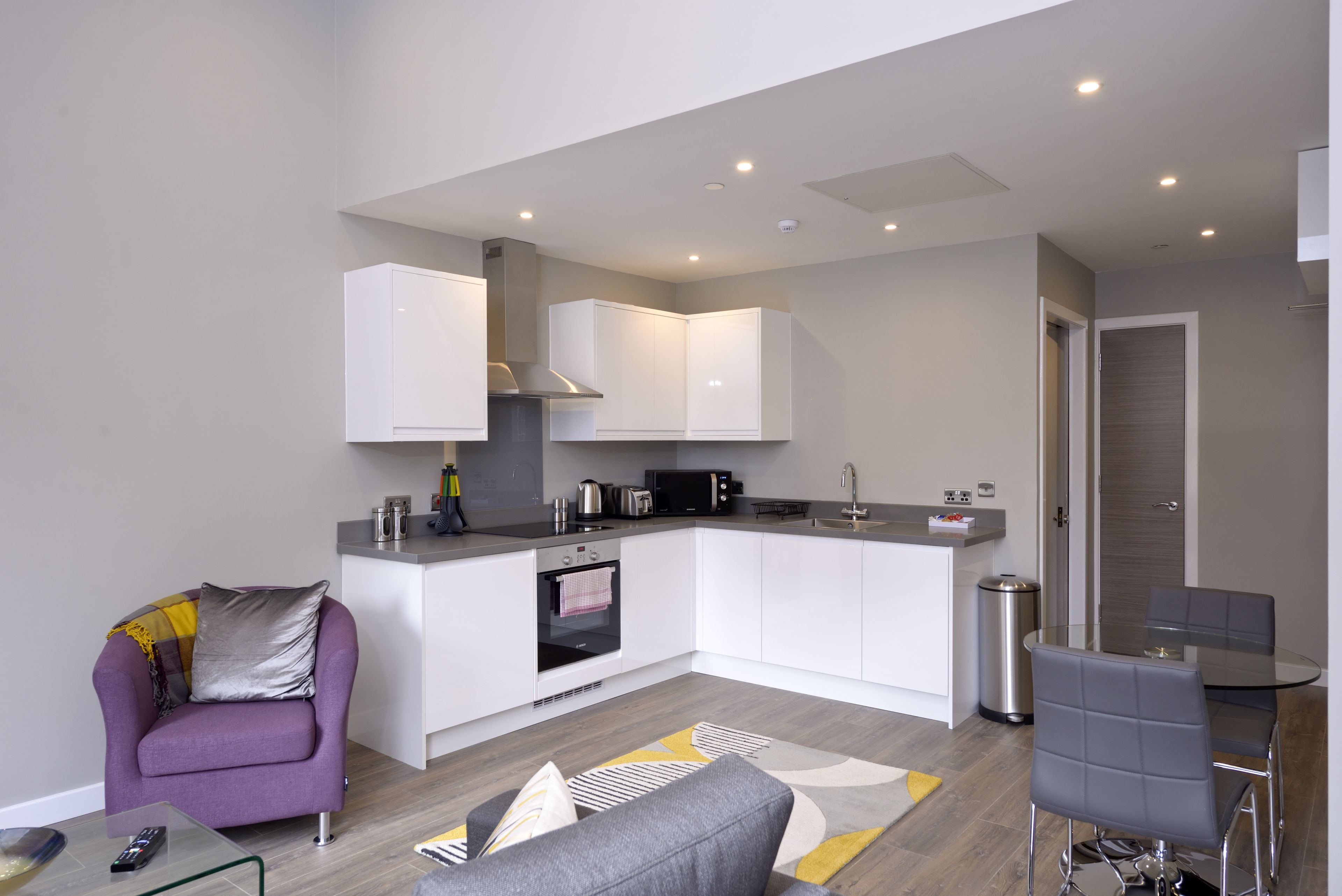 A modern kitchen with white cabinets, a built-in oven, and a stainless steel sink. There is a dining area with a glass table and grey chairs.