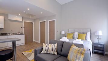 A modern hotel room with a grey sofa, a neatly made bed with yellow and white pillows, a small bedside table with a lamp, and a kitchen area with a sink and cabinets.
