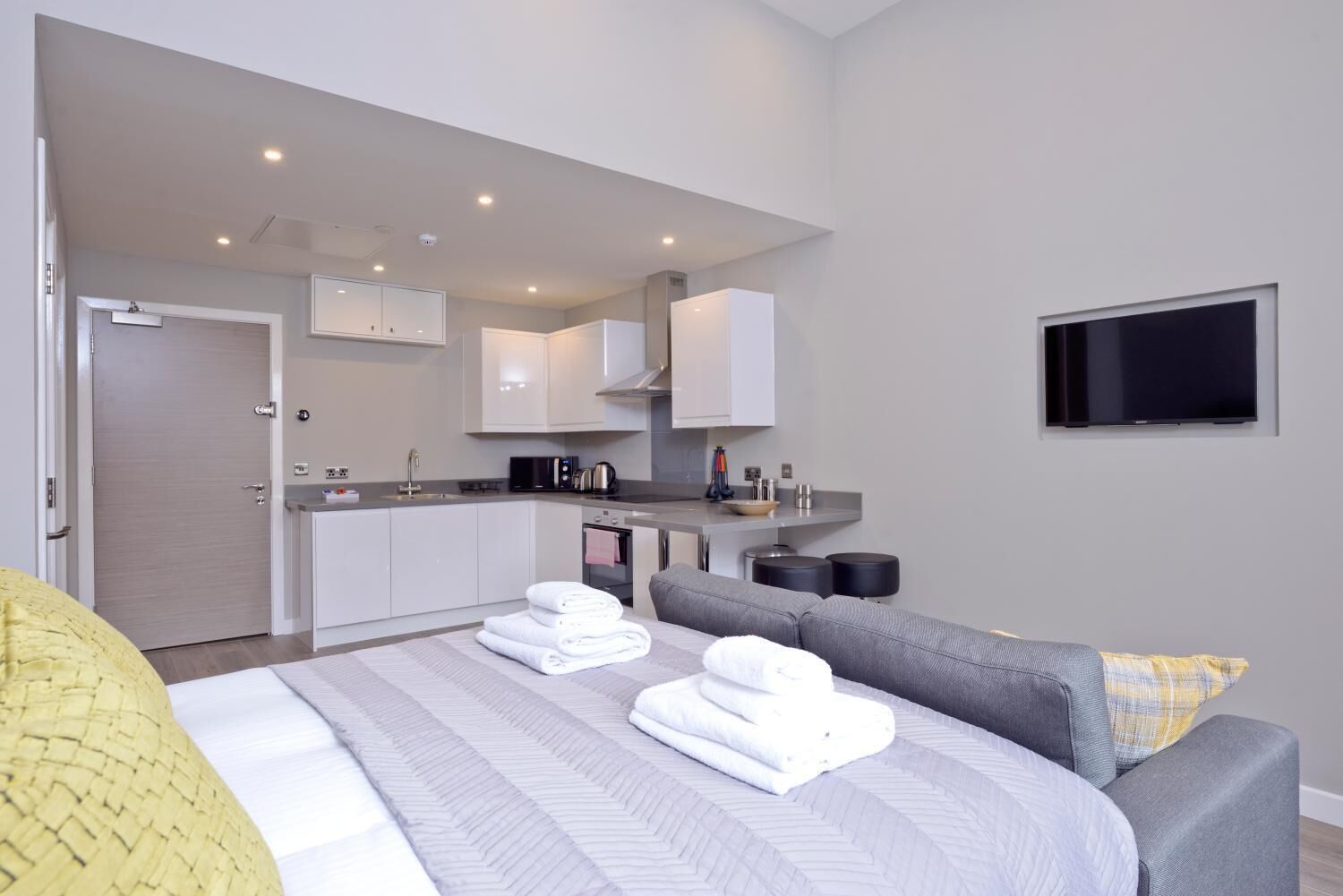 Ein modernes Hotelzimmer mit Bett, grauem Sofa, Fernseher, Küchenzeile und einer Tür.