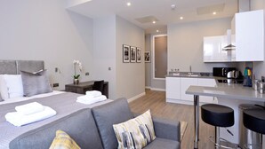 A modern hotel room with a bed, grey sofa, kitchenette, and a small dining area.