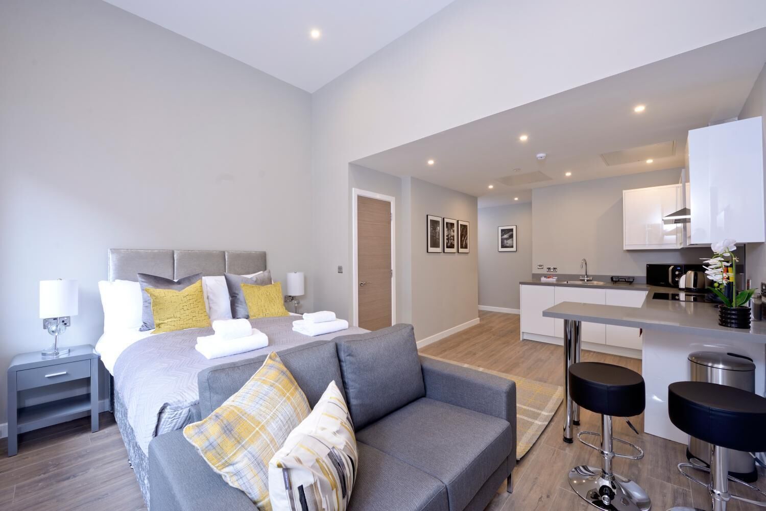 A modern hotel room with a bed, a grey sofa, a small kitchen area, and a seating area with stools.