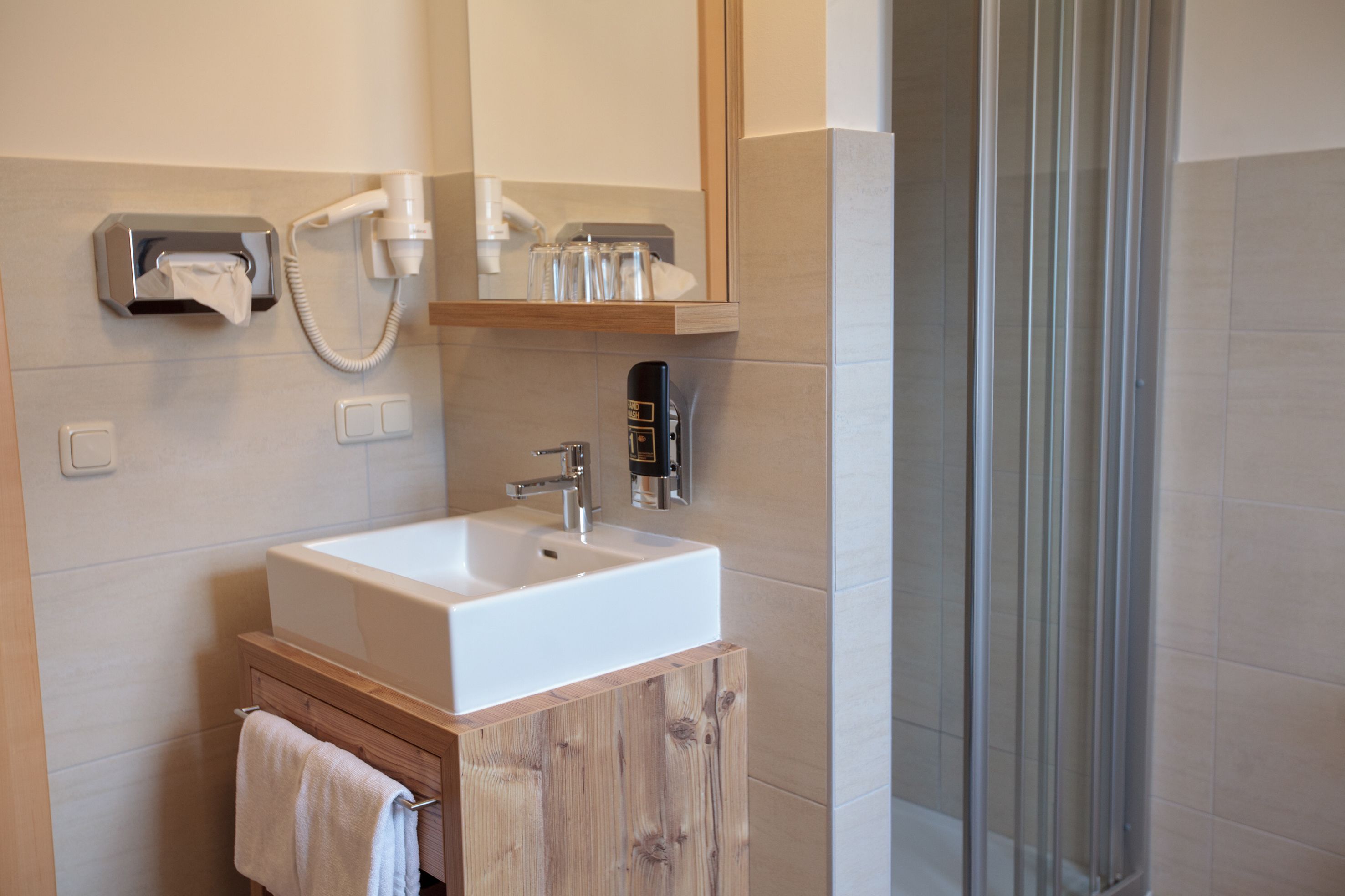 A bathroom with a white sink, a wooden cabinet, a towel hanging on the cabinet, a mirror above the sink, and a shower area with glass doors.