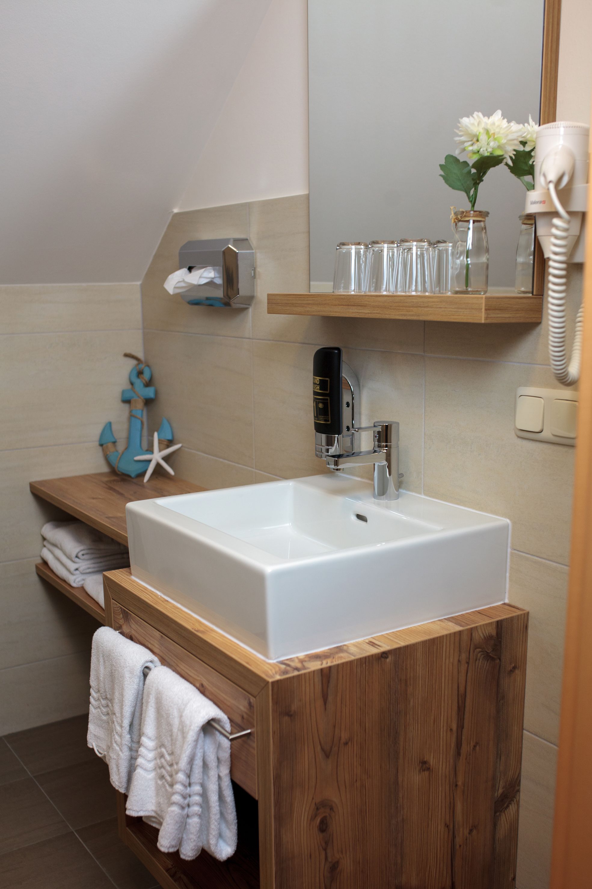A bathroom with a wooden vanity, a white sink, and a mirror above it. There are towels hanging on the side, a small shelf with a jar of flowers, and a hairdryer on the wall.