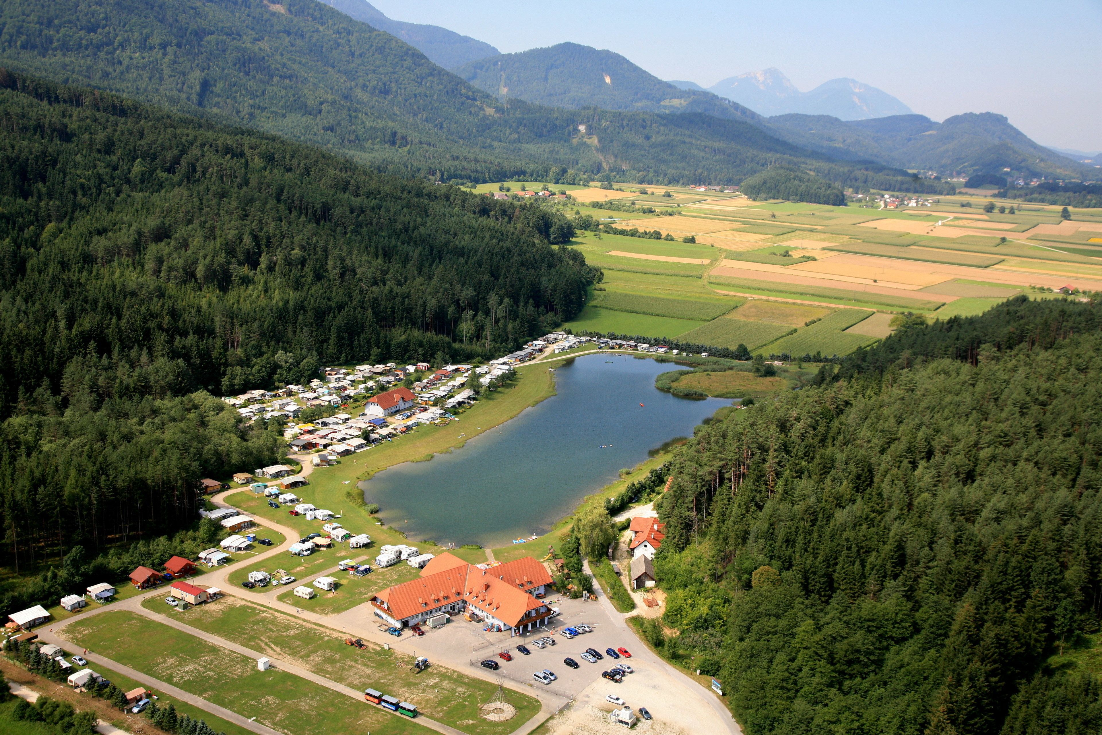 An aerial view of a lakeside campground with RVs and cabins, surrounded by forested hills and fields.