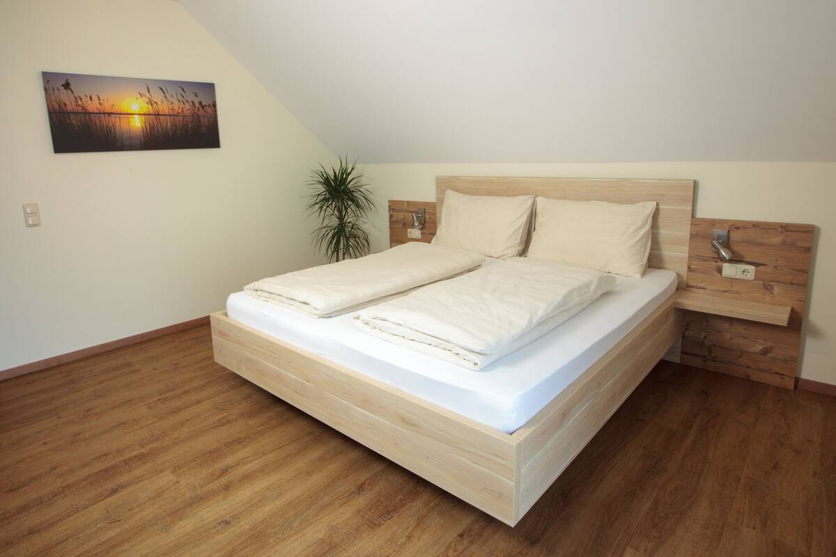 Een moderne slaapkamer met een houten bed, een nachtkastje, een wandcontactdoos en een groot schilderij van een zonsondergang.