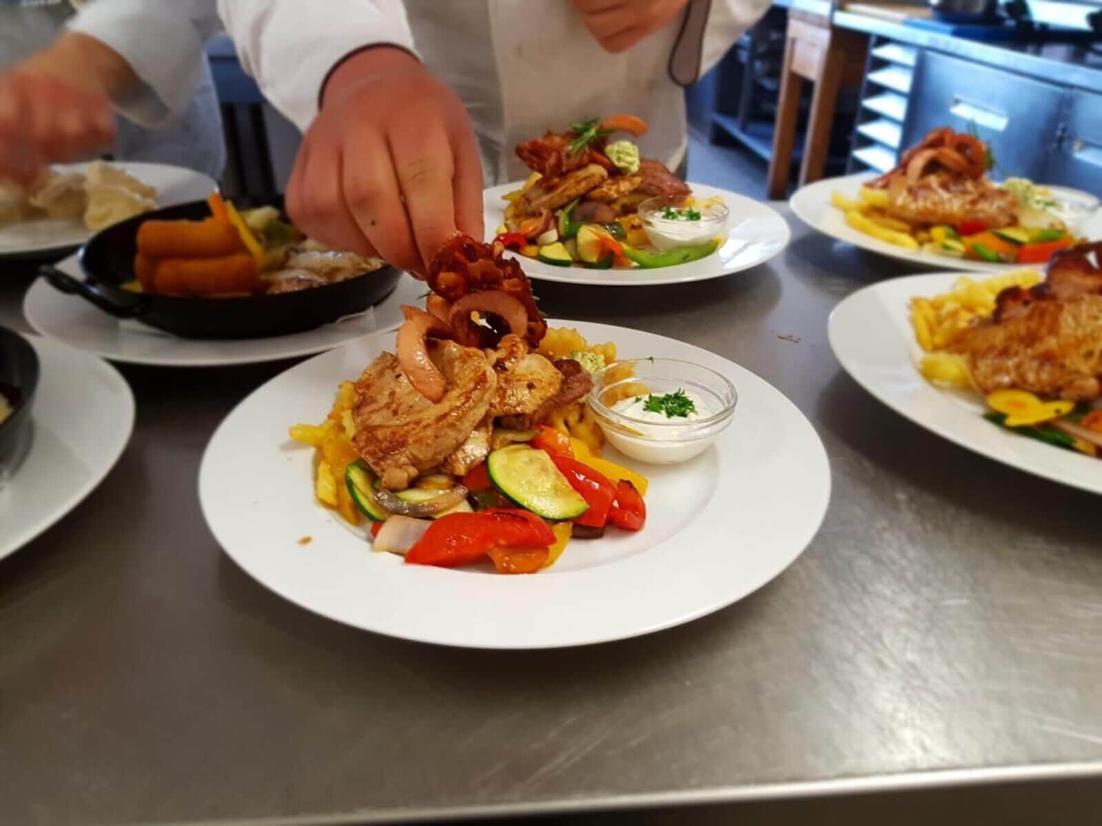 A chef preparing a dish with grilled chicken, vegetables, and a side of sauce on a stainless steel table.