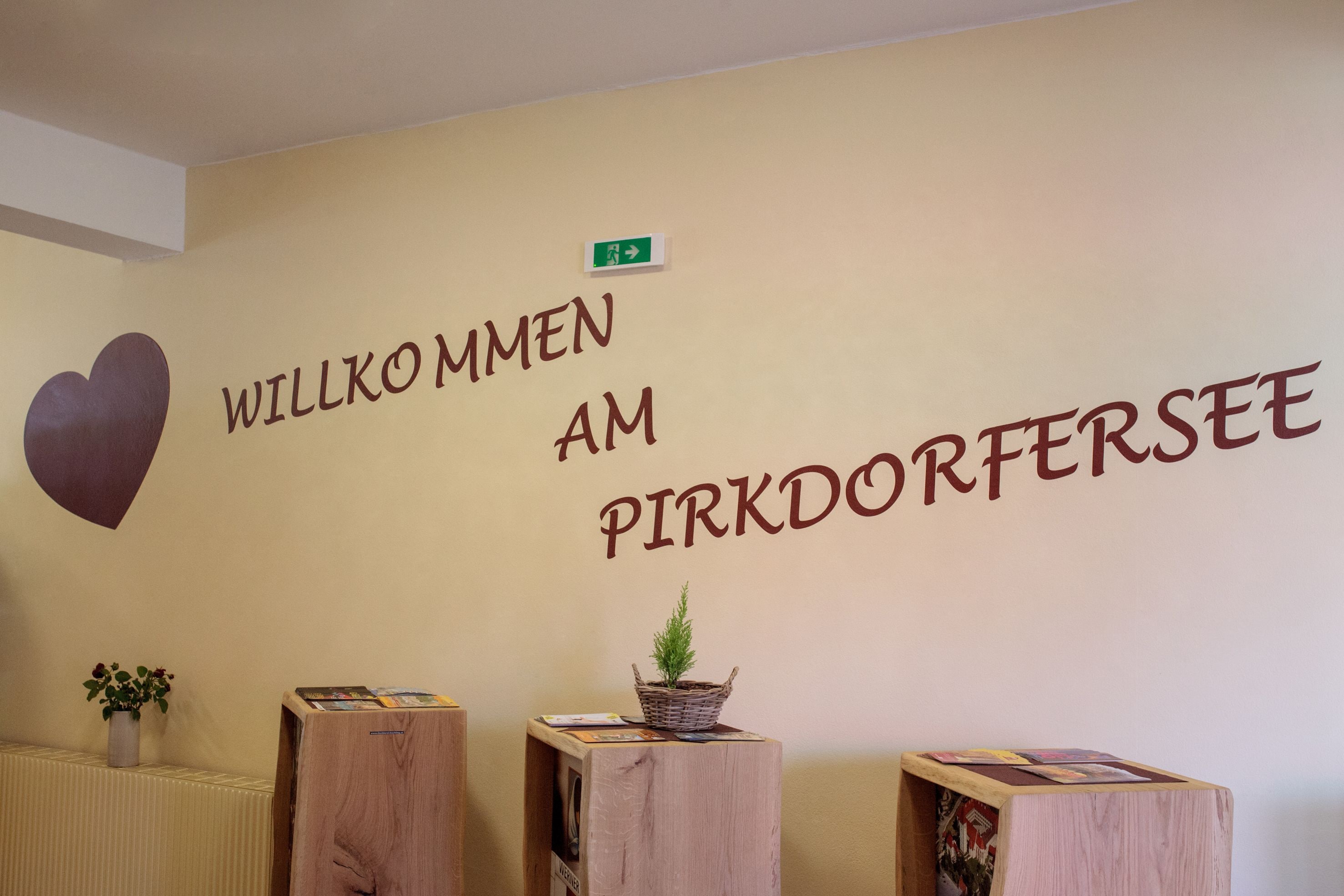 A reception area with a wall sign that reads "Willkommen am Pirkdorfersee" and a heart symbol. There are wooden stands with magazines and a small plant.