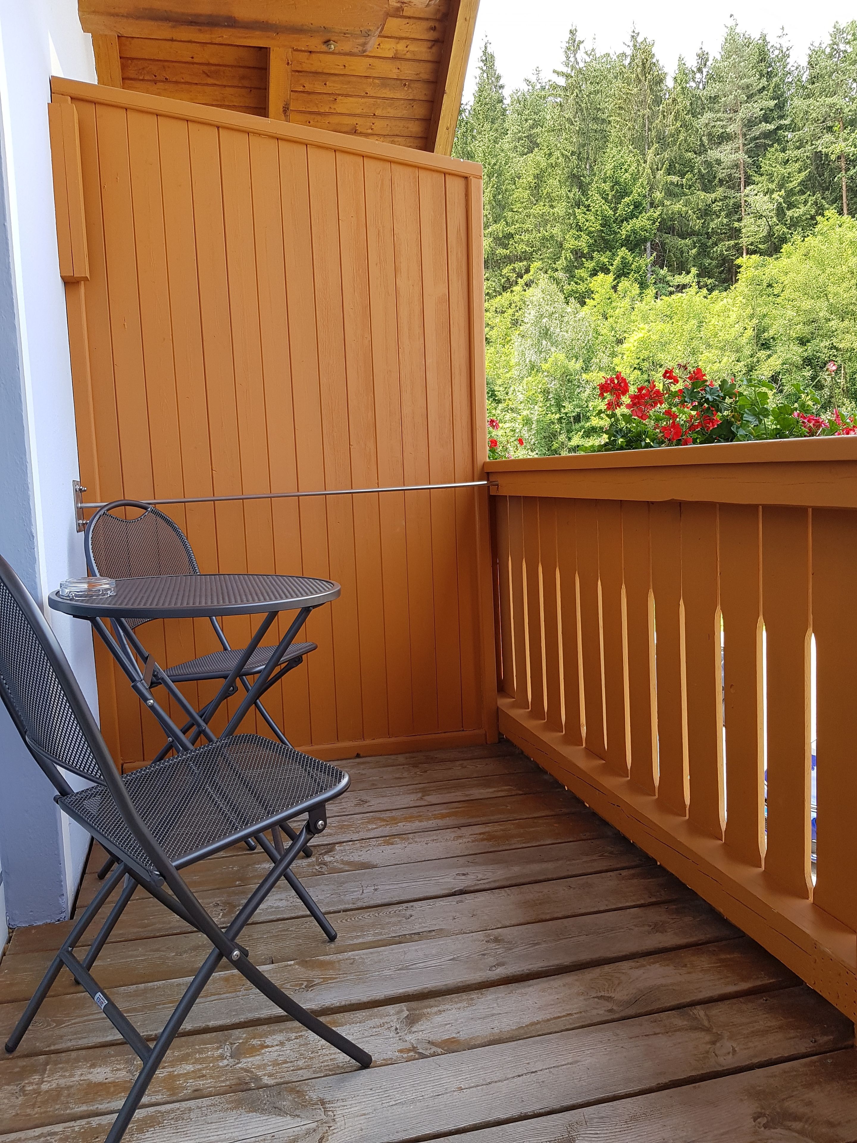 Un deck de madera con una mesa y sillas de metal, rodeado por un cerco de madera y vistas al follaje.
