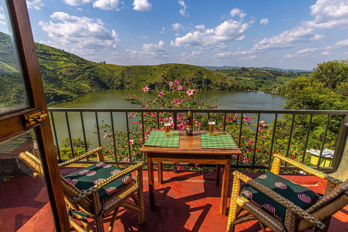 A balcony with wicker chairs and a table, overlooking a lake and hills.