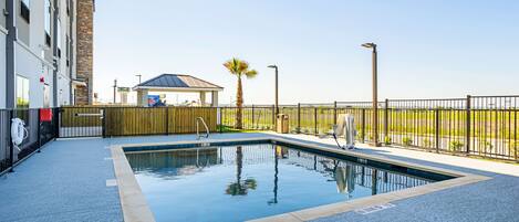 A swimming pool with a tiled deck, a wooden fence, and a building in the background.