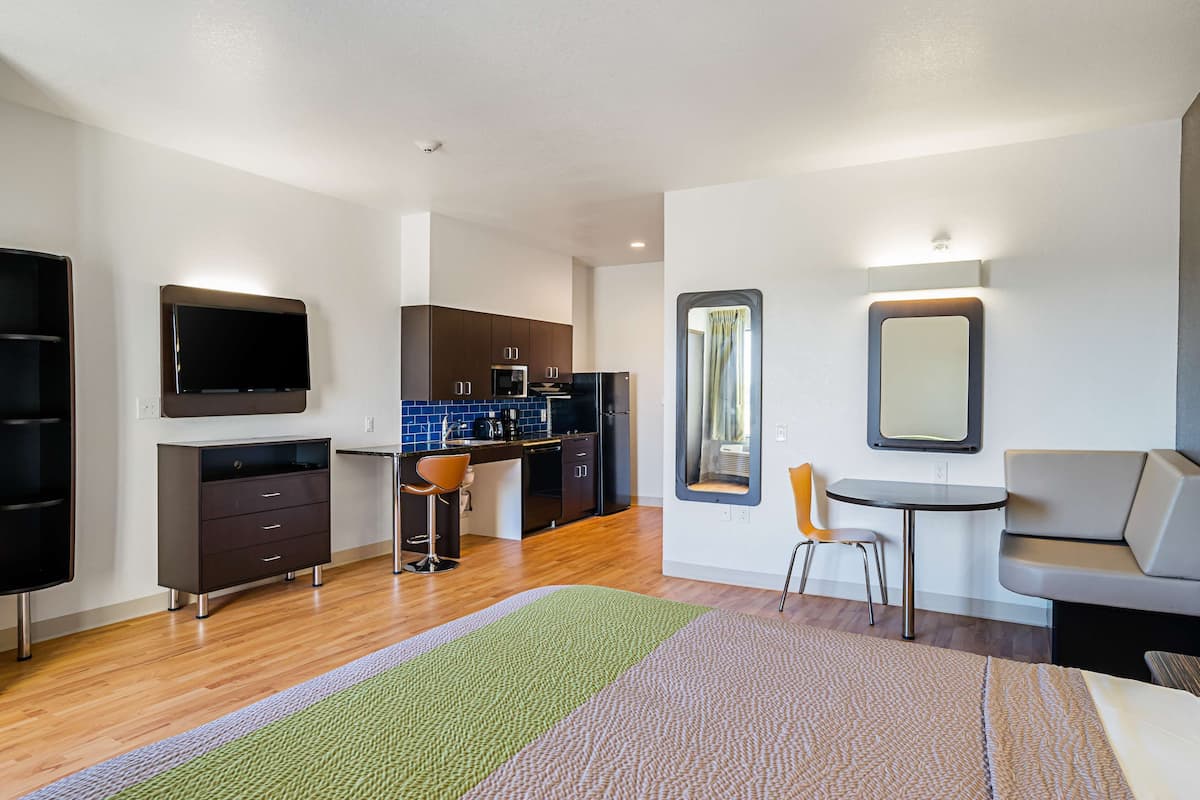 Una habitación de hotel moderna con kitchenette, televisor de pantalla plana, zona de estar con mesa y sillas, y un espejo grande.