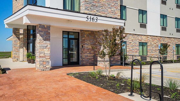 A modern building with a stone facade, numbered 5163, featuring a covered entrance and a parking area with a metal bike rack. - Studio 6 Buda, TX (Buda)