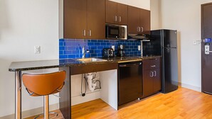 A compact kitchen with brown cabinets, a black refrigerator, a microwave, and a sink with blue tiles. - Studio 6 Buda, TX (Buda)