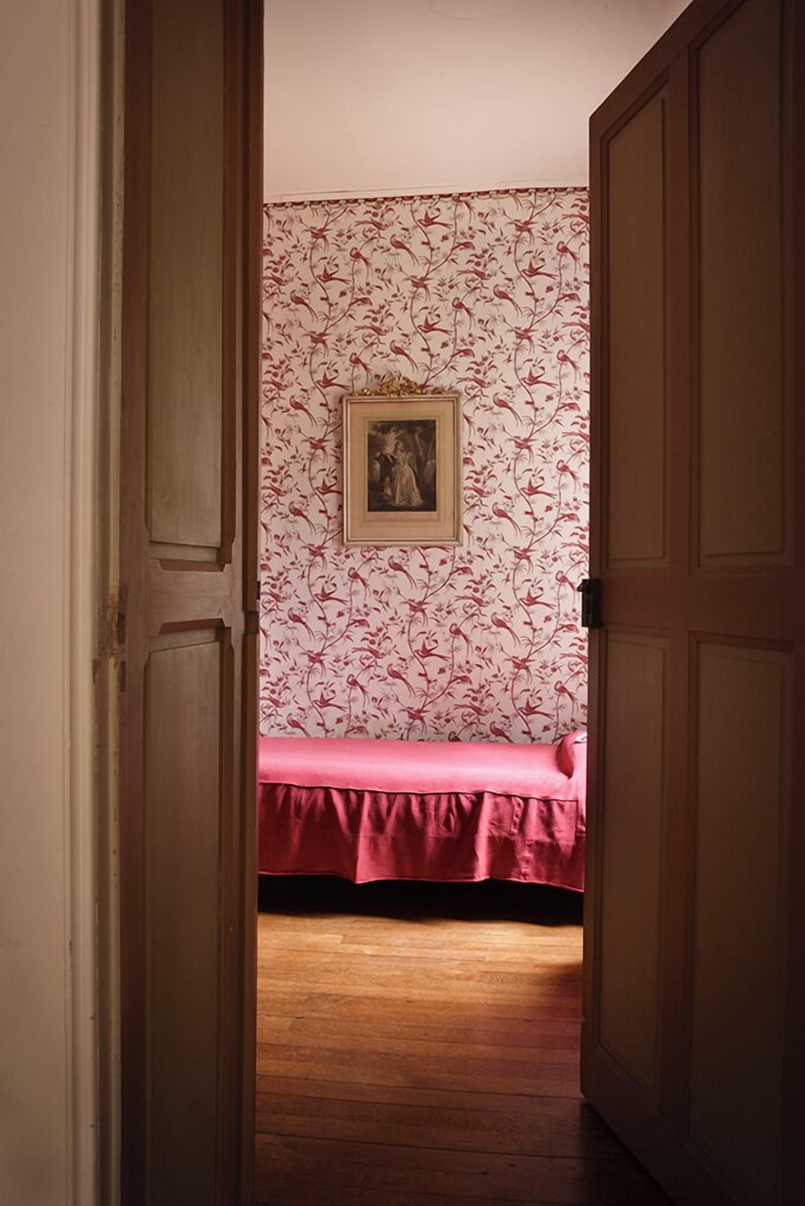 Une pièce avec un plancher en bois, un lit recouvert d’une courtepointe rose et un papier peint à motifs floraux.