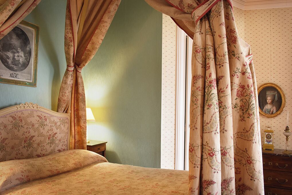 Un dormitorio con cama con dosel, cortinas florales, un cuadro enmarcado en la pared, un aparador con un reloj y una mesita con una lámpara.