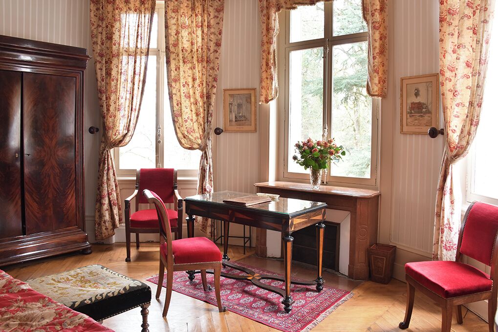 A traditional room with a wooden cabinet, a red upholstered chair, a glass table, and a window with floral curtains.