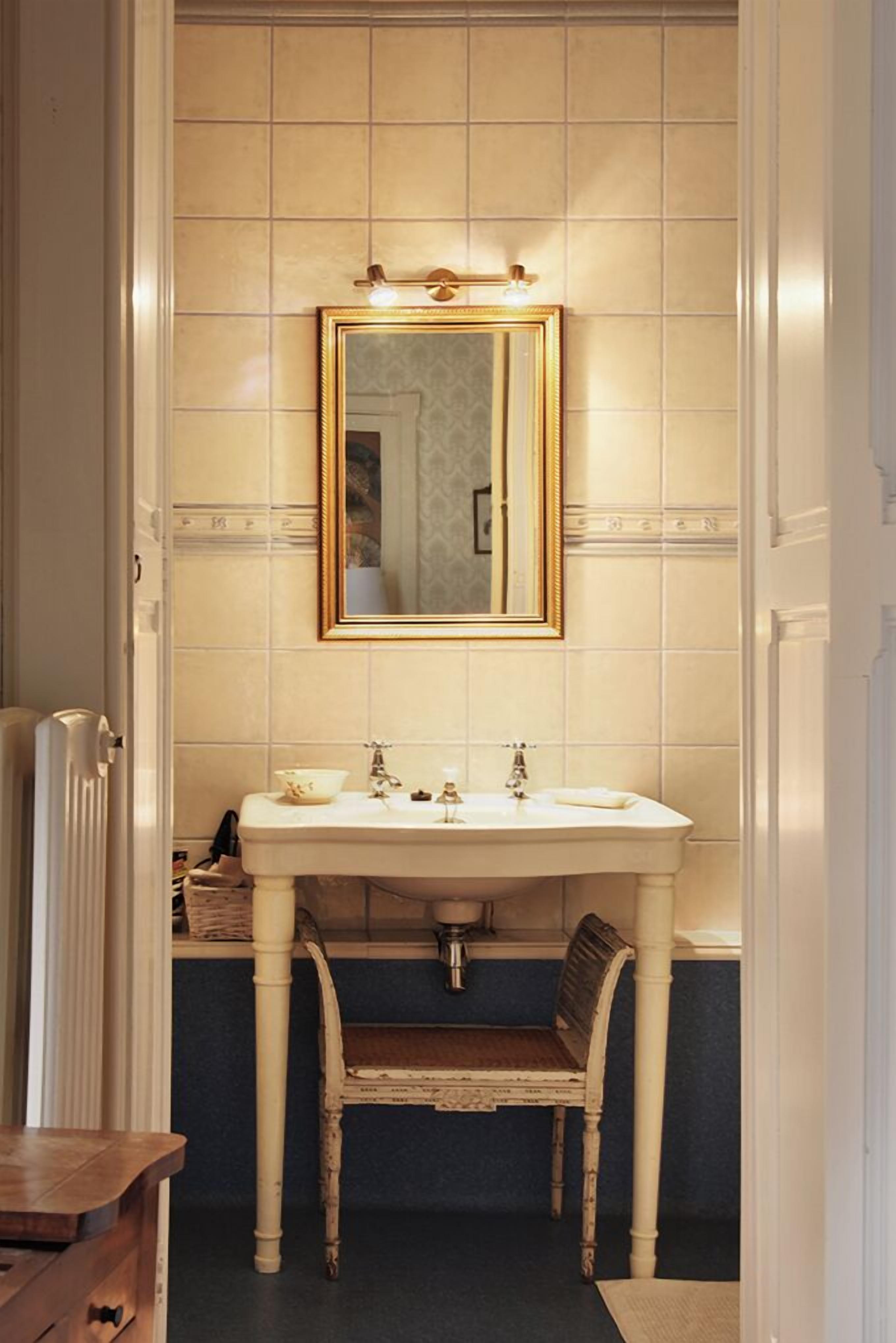 Een badkamer met een witte wastafel, een spiegel met gouden lijst en een stoel.