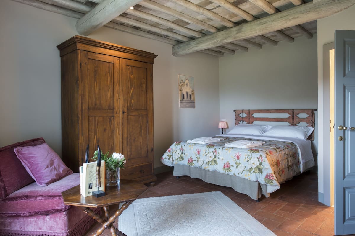 A bedroom with a wooden wardrobe, a bed with floral bedding, a pink lamp, and a red sofa.