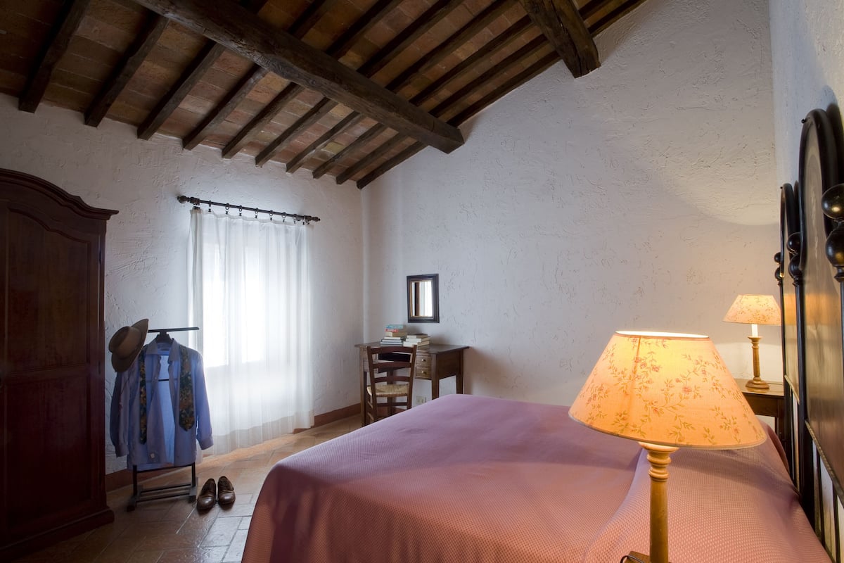 A bedroom with a wooden ceiling, a bed with a pink bedspread, a desk with a chair, a wardrobe, and a window with curtains.