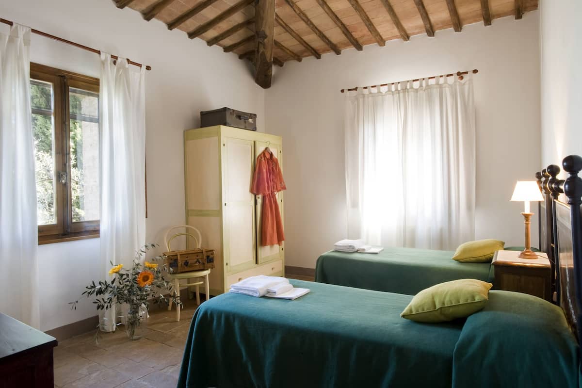 A room with two beds, a wooden ceiling, a cabinet, a lamp, and a window with curtains.