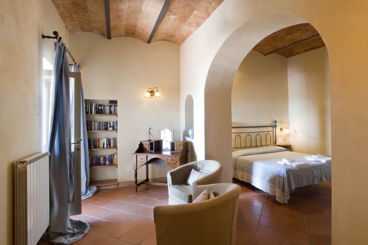 A bedroom with a bed, a chair, a desk with a lamp, a bookshelf, and a window with curtains.