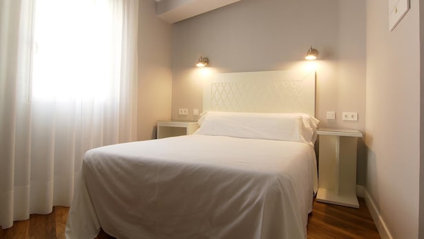 A neatly made bed in a room with a white headboard, two bedside lamps, and a window with sheer curtains. - Hostal los Castañuelos (Candeleda)