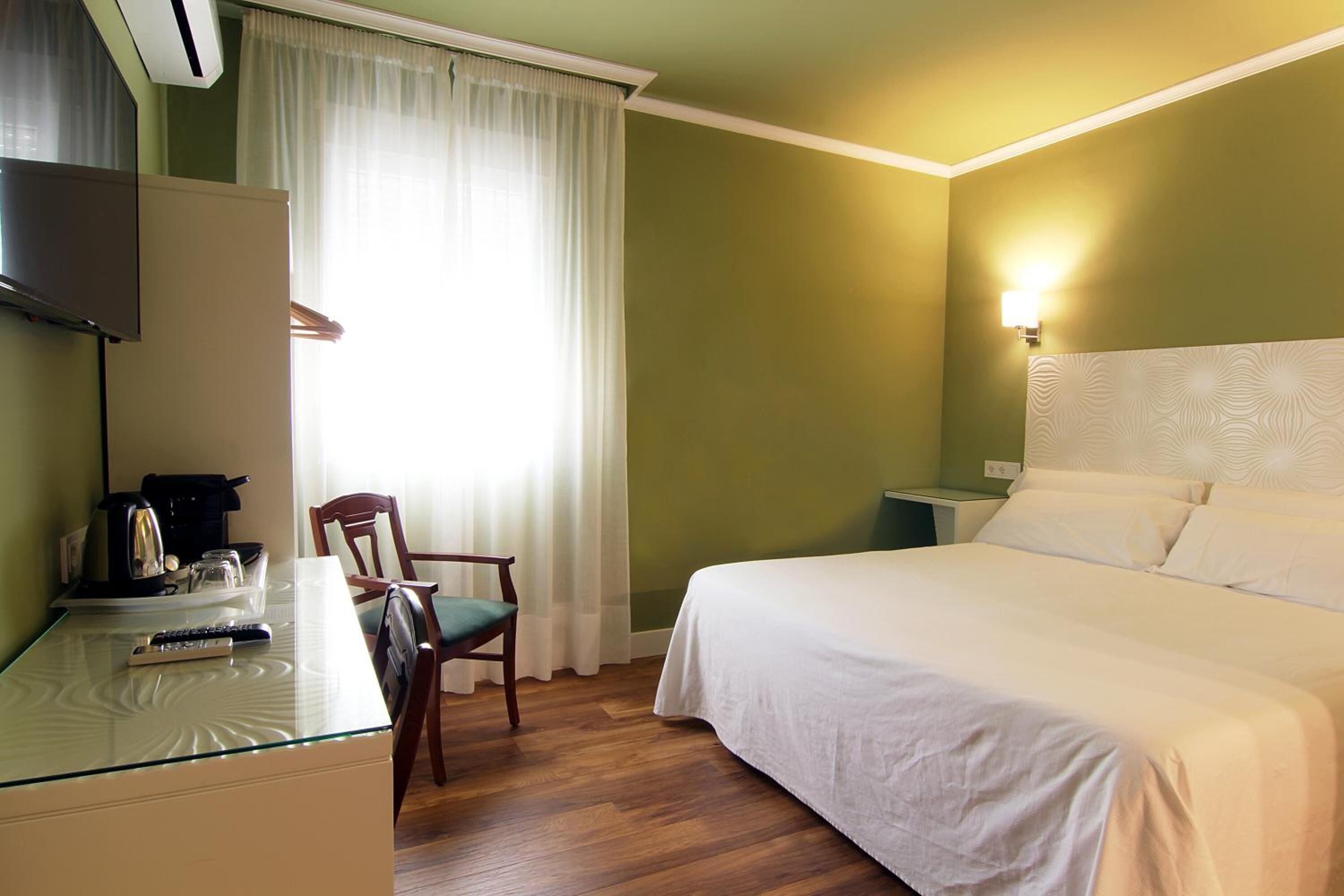 A hotel room with a bed, a desk with a remote control, a chair, a television, and a window with curtains.
