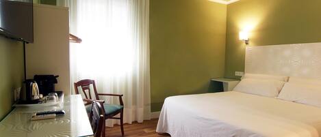 A hotel room with a bed, a desk with a remote control, a chair, a television, and a window with curtains.
