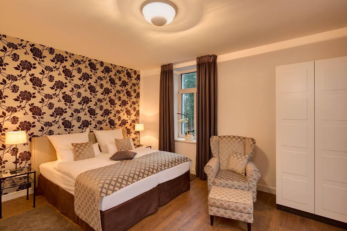 A bedroom with a large bed, a chair, a nightstand, a window with curtains, and a wardrobe.