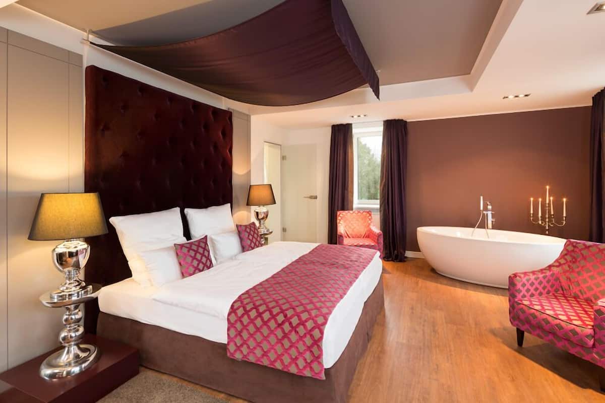 A modern hotel room with a large bed, a sitting area with a chair, a bathtub, and a window with curtains.