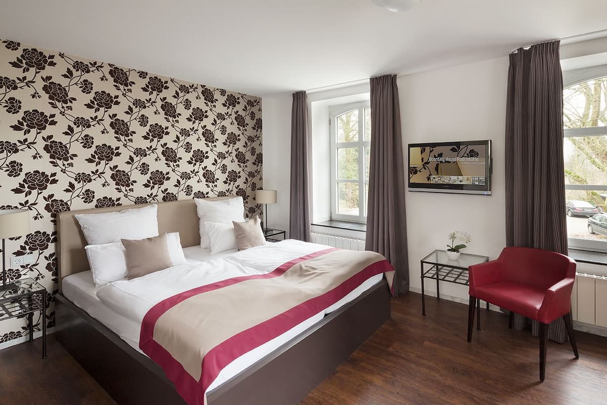 A neatly arranged bedroom with a large bed, a small table, a chair, and a window with curtains.