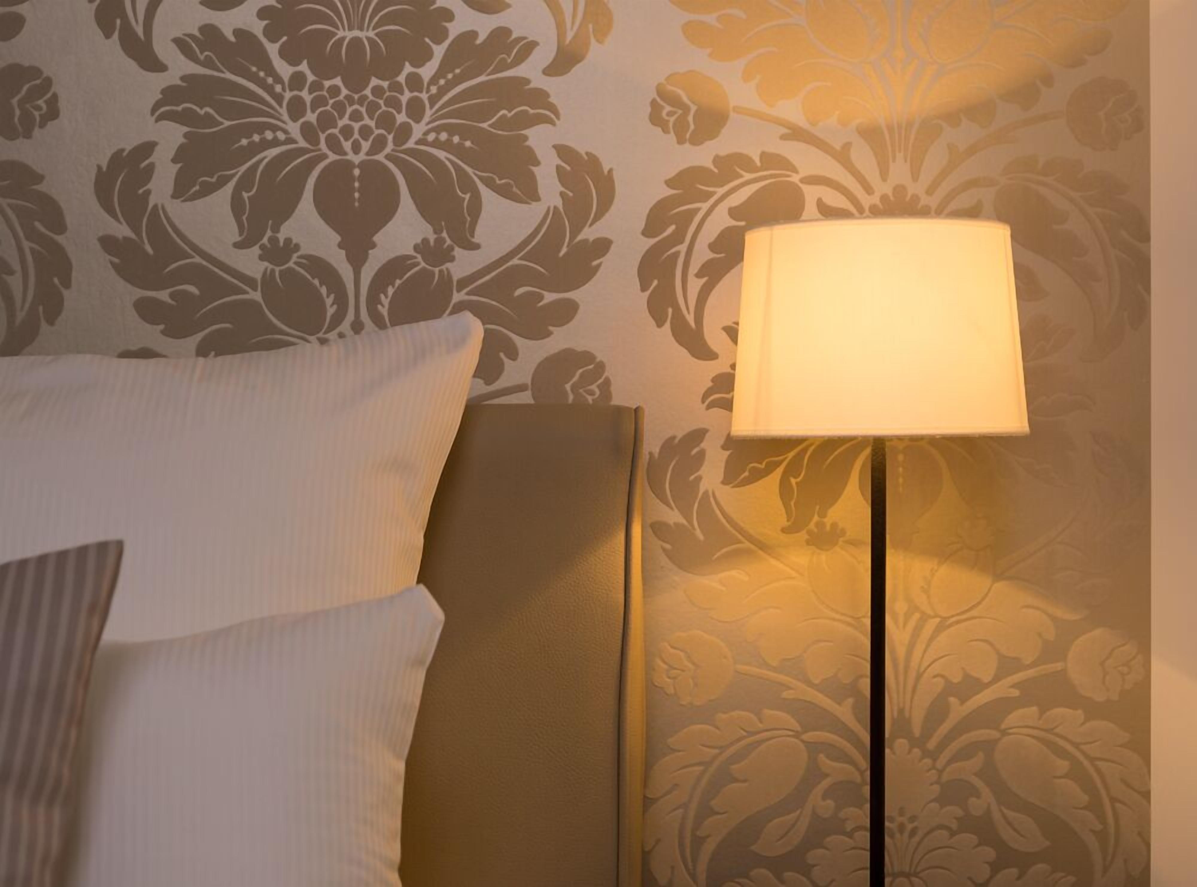 Una cama bien hecha con sábanas blancas y una sola almohada, una mesita de noche con lámpara, y una pared con estampado floral.