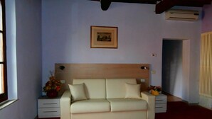 A bedroom with a beige sofa, a framed picture on the wall, a window, and an air conditioning unit. - Bed and Breakfast La Torretta (Cascina)