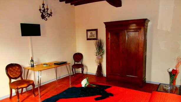 A room with a wooden floor, a desk with a chair, a bed with a red and black blanket, a wooden wardrobe, a chandelier, and a vase with flowers. - Bed and Breakfast La Torretta (Cascina)