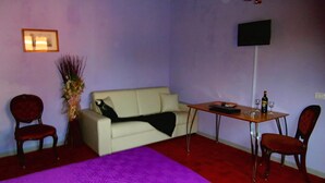 A room with a purple carpet, a white sofa, a wooden table with a bottle and glasses, and a TV on the wall. - Bed and Breakfast La Torretta (Cascina)
