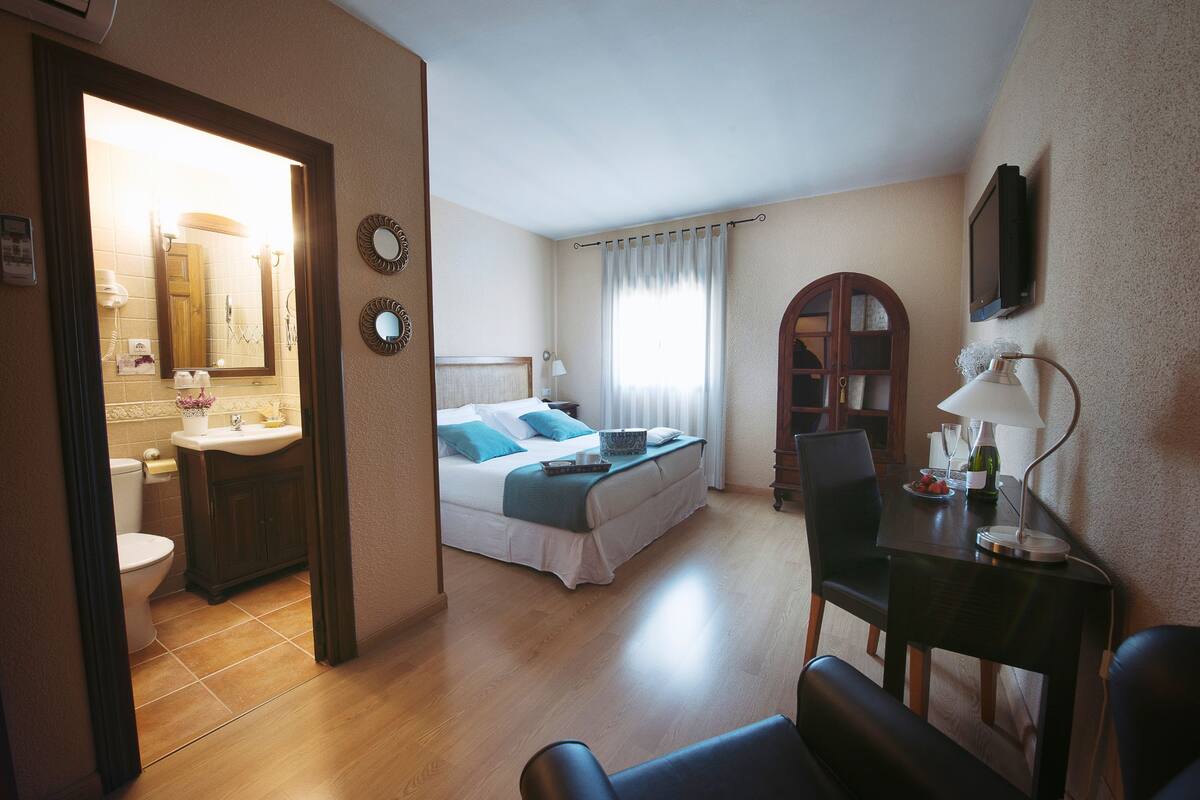 Quarto de hotel com cama, escrivaninha, cadeira, televisão, janela com cortinas e banheiro com vaso sanitário e pia.